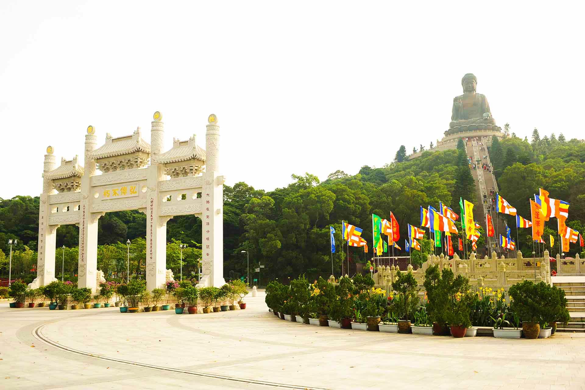 Tian Tan Buddha statue on hilltop with traditional Chinese gateway arch and colorful international flags at Po Lin Monastery,